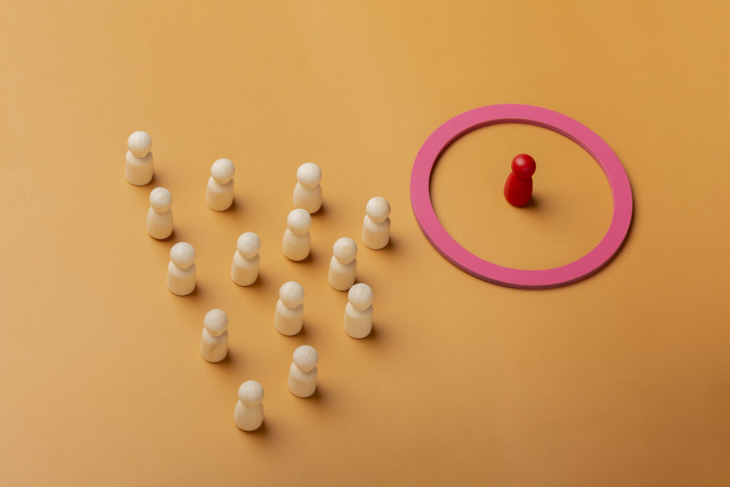 A imagem mostra uma peça vermelha solitária posicionada dentro de um círculo rosa, separada de um grupo de várias peças de madeira de cor clara, todas agrupadas ao fundo. O fundo é de cor laranja, criando um contraste com as peças. A cena sugere conceitos como segmentação, exclusão, individualidade, ou direcionamento de grupo, temas que podem estar relacionados a marketing, segmentação de público, ou gestão de leads em CRM.