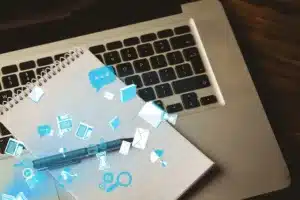 Imagem de um caderno e caneta sobre um laptop, com ícones de comunicação digital flutuando, simbolizando produtividade e tecnologia.