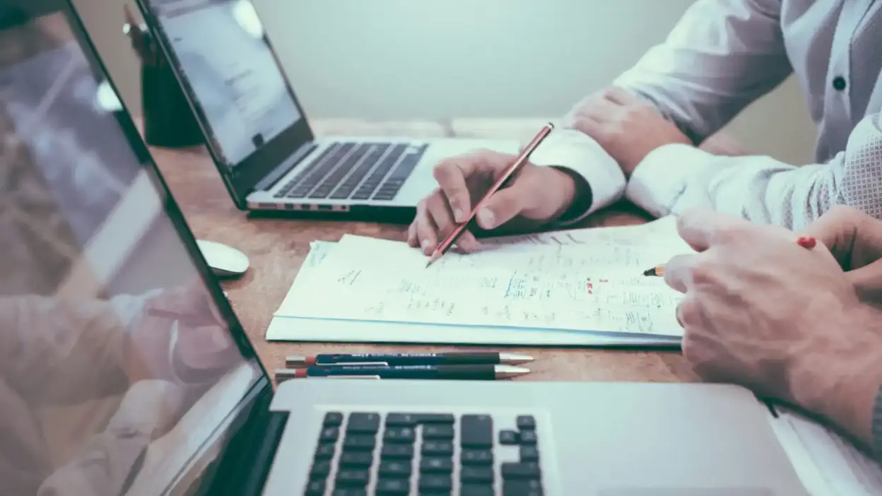Pessoa trabalhando em computador com notebooks, tablets e documentos, demonstrando processo de automatização de checklists e relatórios em 4 passos.