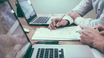 Pessoa trabalhando em computador com notebooks, tablets e documentos, demonstrando processo de automatização de checklists e relatórios em 4 passos.