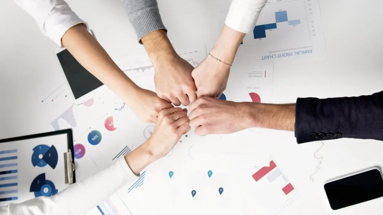 Equipe de negócios unindo as mãos em sinal de trabalho em equipe, com gráficos e documentos em uma mesa de escritório, simbolizando gestão moderna, desafios e estratégias empresariais.