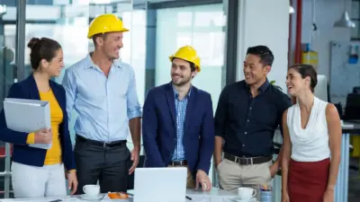 Equipe de engenheiros e profissionais de construção civil sorrindo e conversando durante reunião no escritório, com equipamentos de segurança como capacetes.