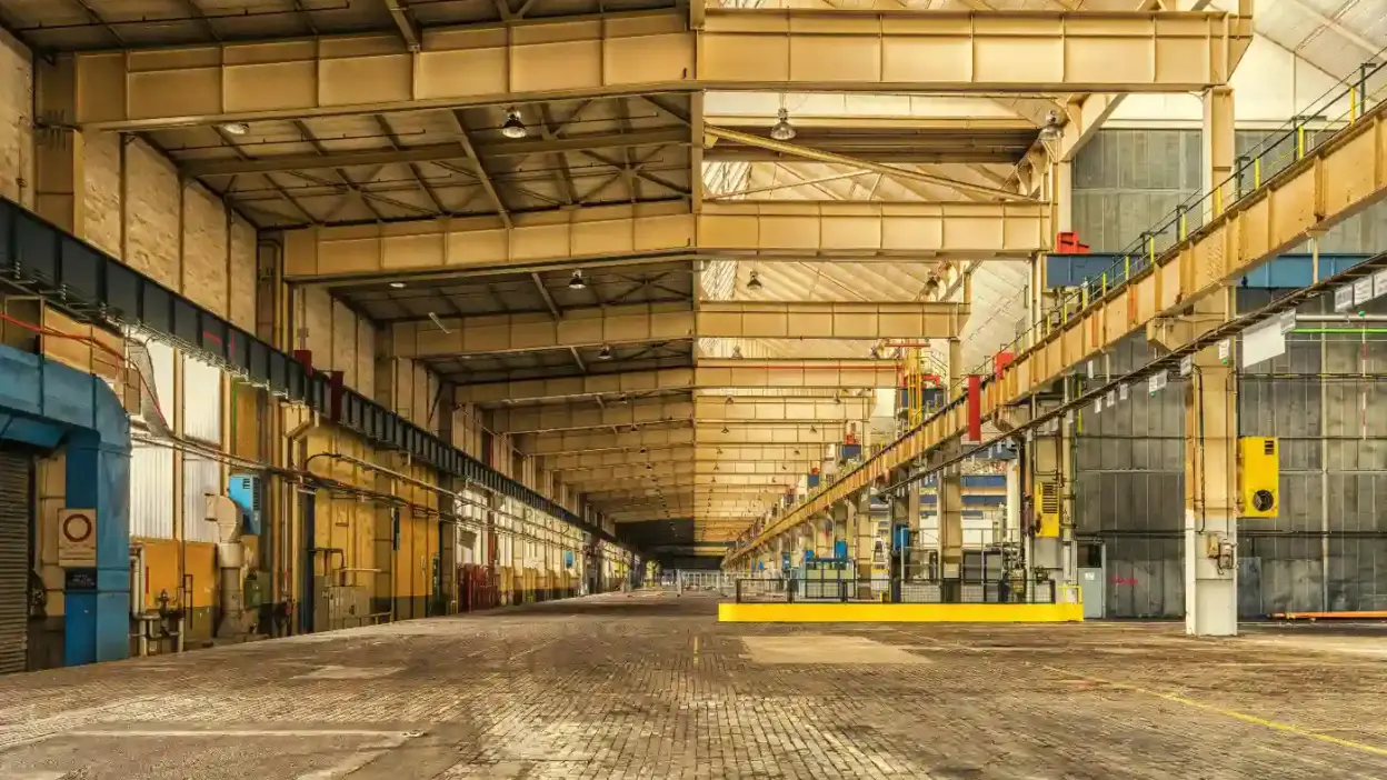 Imagem de um grande galpão industrial vazio, com teto alto, estruturas metálicas, piso de concreto e áreas organizadas para operações industriais.