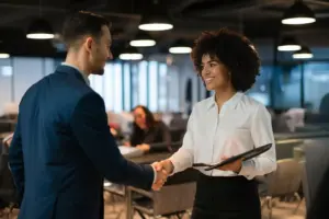 Homem e mulher em reunião de negócios trocando um aperto de mãos em ambiente de escritório moderno, sorrindo e transmitindo profissionalismo.