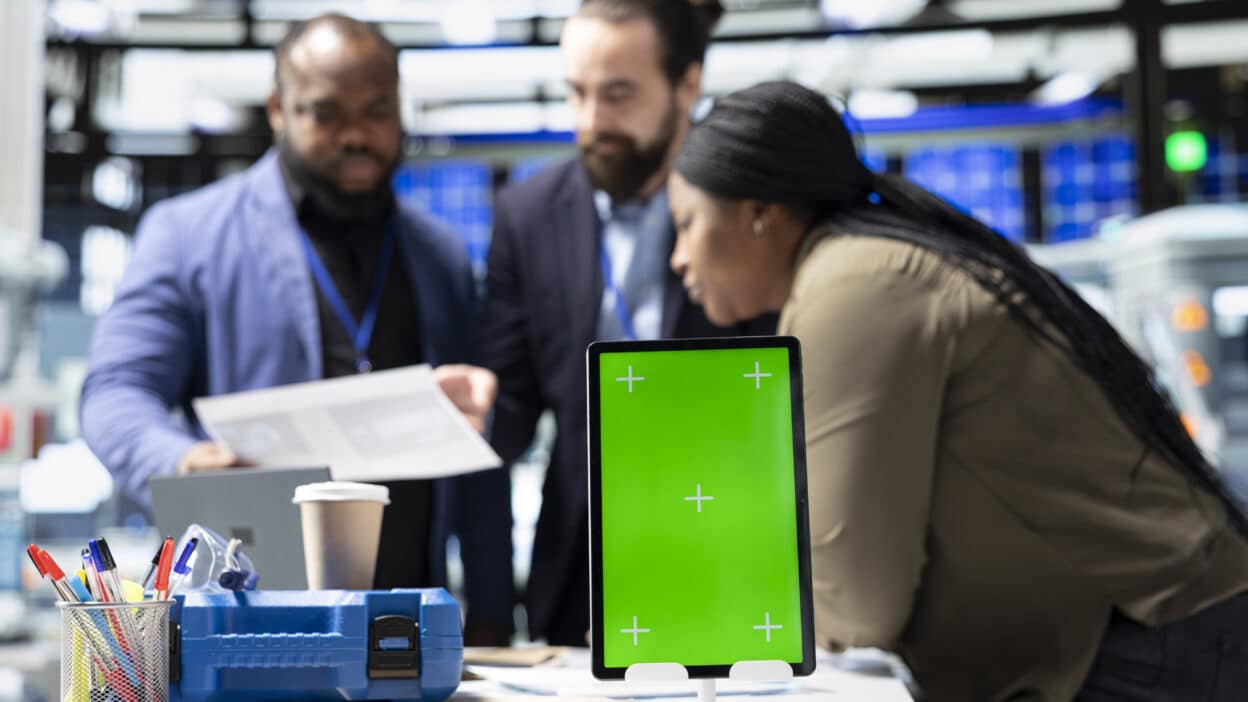 Equipe de profissionais reunida em ambiente tecnológico analisando documentos e equipamentos, com tablet sobre a mesa durante discussão de projeto.