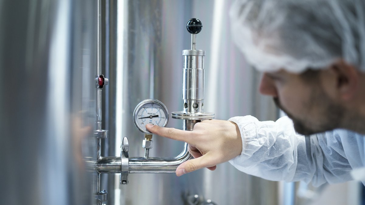 NR-13 para caldeiras: o que a norma exige do comprador 2 Técnico de uniforme branco aponta para um manômetro instalado em tubulação de aço inox