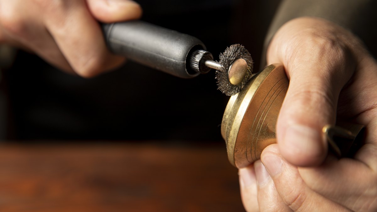 Para que serve a micro retífica e como usar com segurança 2 Mão segurando peça redonda de latão enquanto outra mão aproxima uma micro retífica com roda de polimento