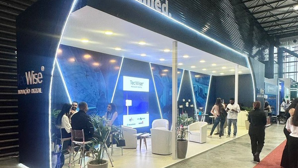 O que é cenografia: como funciona em eventos e stands 3 Stand com fachada azul-escura contornada por linhas de LED branco, poltronas brancas, televisores e visitantes no corredor da feira