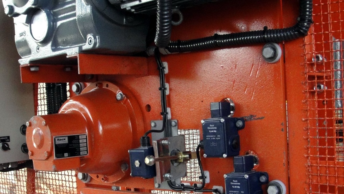 O que é elevador cremalheira e como funciona 2 Detalhe do acionamento de elevador cremalheira: motor, freio e chaves elétricas montados na estrutura laranja da cabina