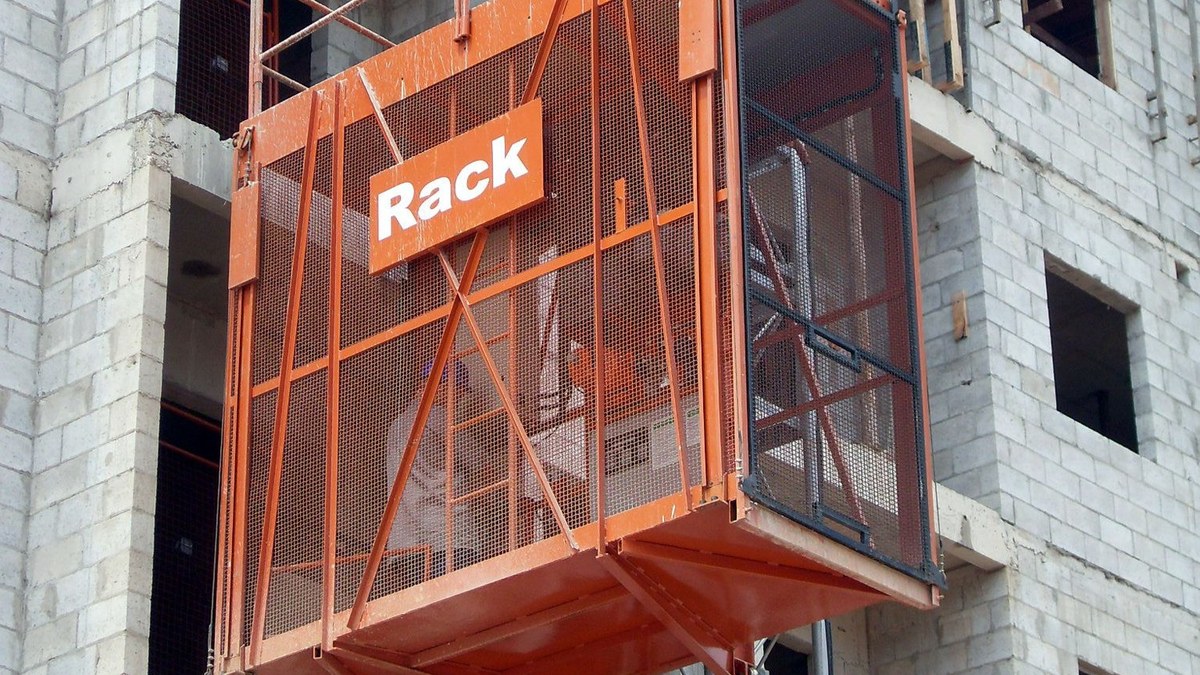 Elevador cremalheira e NR-18: o que a norma exige 2 Cabina laranja de elevador cremalheira com a marca Rack parada junto a um pavimento de prédio de concreto em construção