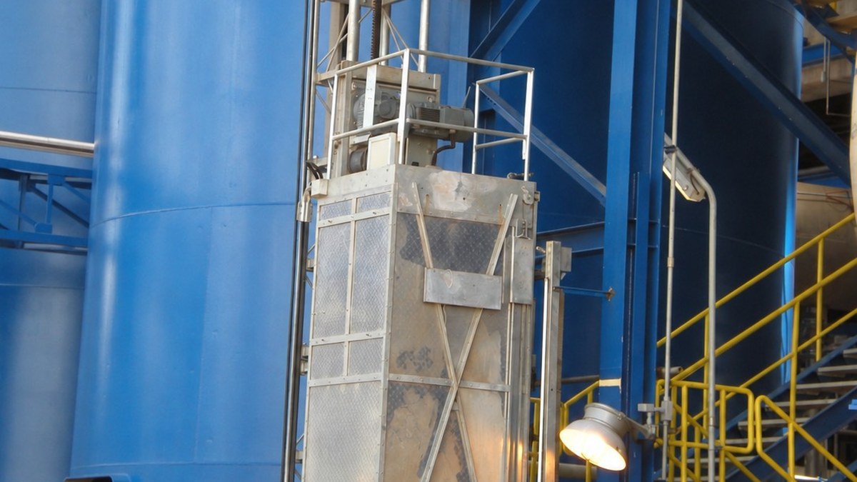 O que é elevador cremalheira e como funciona 3 Elevador cremalheira com cabina metálica cinza instalado ao lado de estrutura industrial azul e escadas amarelas