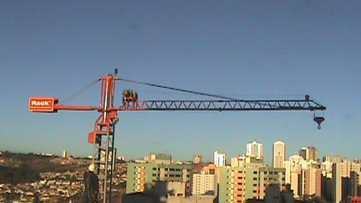Tipos de elevador de obra e quando usar cada um 2 Grua de pequeno porte laranja com a marca Rack sobre a laje de um prédio, com lança treliçada estendida sobre a cidade ao entardecer