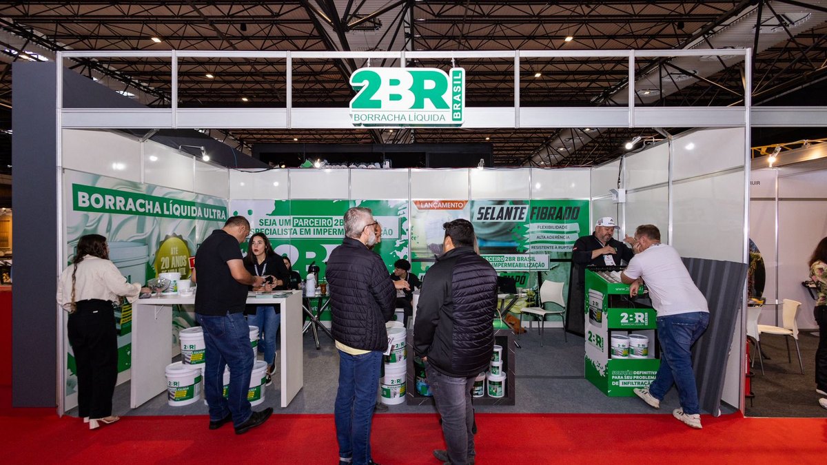 Stand ou estande: o que é e para que serve na feira 2 Stand de painéis brancos com testeira verde, balcões de atendimento e visitantes conversando com a equipe do expositor