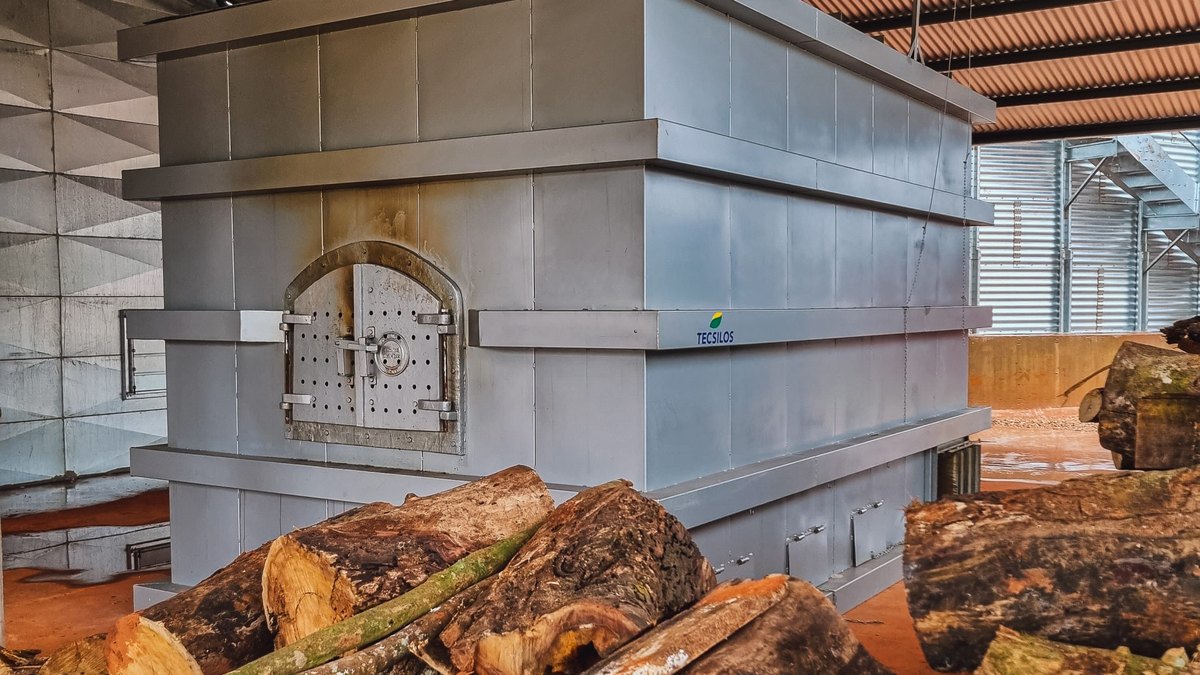 Secagem de grãos: como funciona e quais sistemas existem 2 Fornalha metálica a lenha instalada sob um galpão coberto, com porta de alimentação frontal e toras de madeira empilhadas na frente