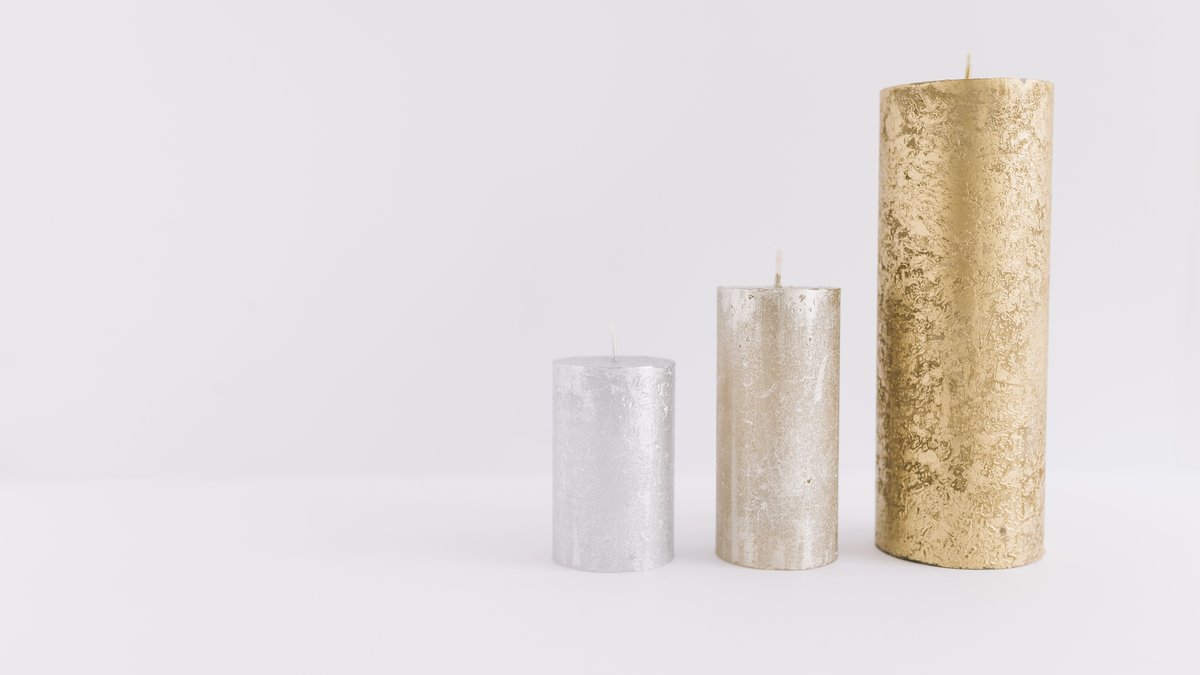 Tipos de vela: maço, 7 dias, rechaud e decorativa 3 Três velas cilíndricas decorativas, duas prateadas e uma dourada, de alturas diferentes sobre fundo branco